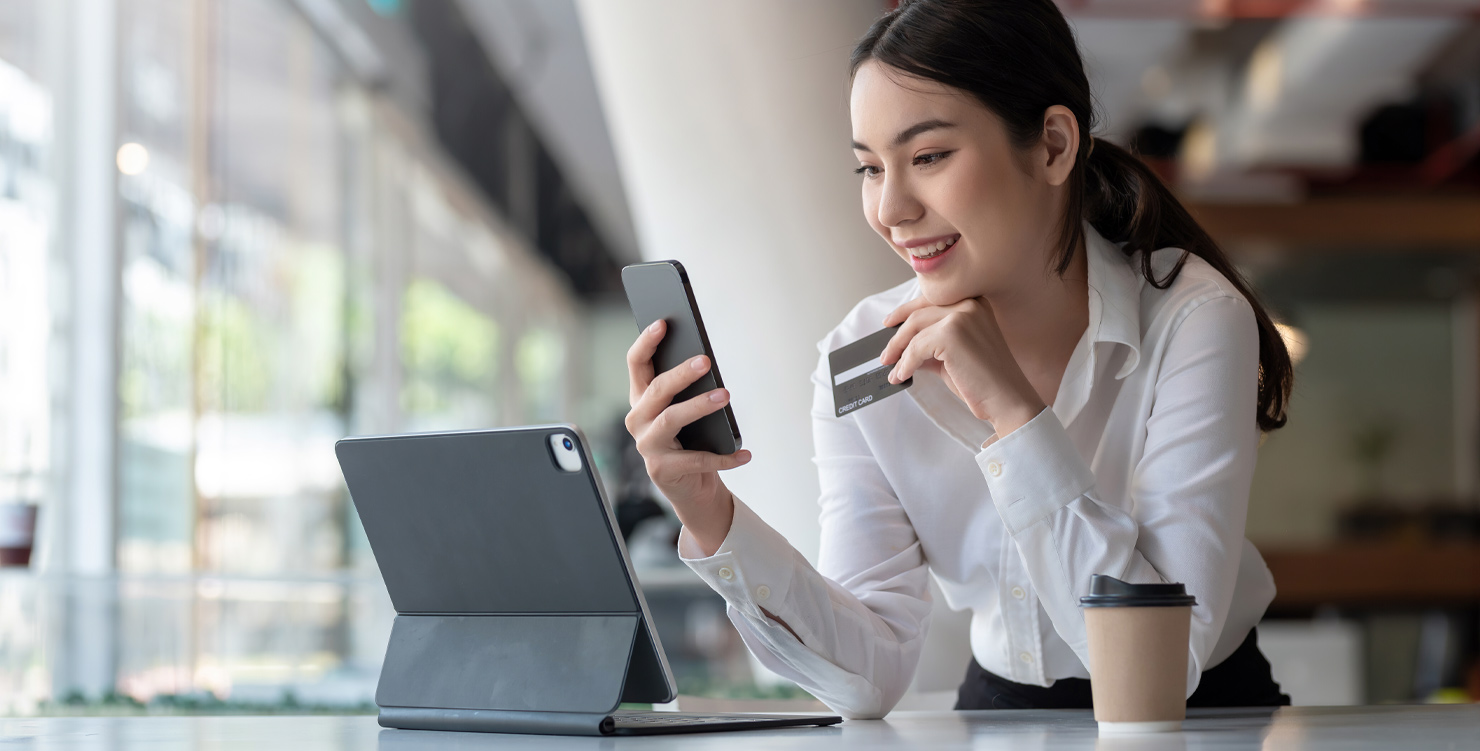 Businesswoman smiling while using a smartphone and credit card with a laptop and coffee beside her.