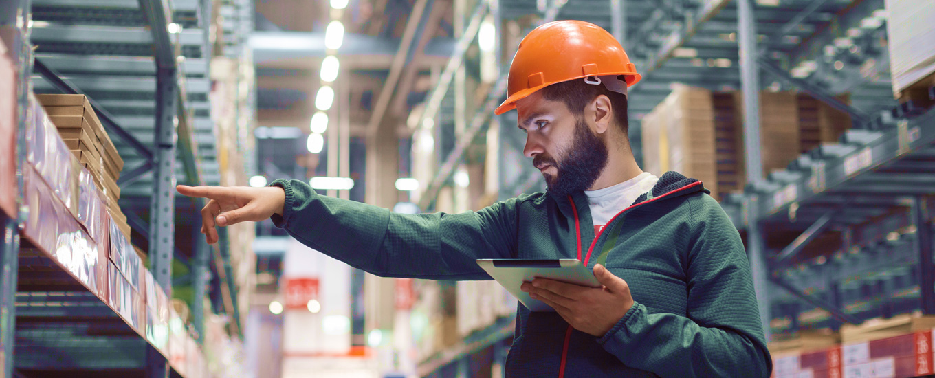 Worker wearing a hard hat using tablet to check inventory in a warehouse aisle.