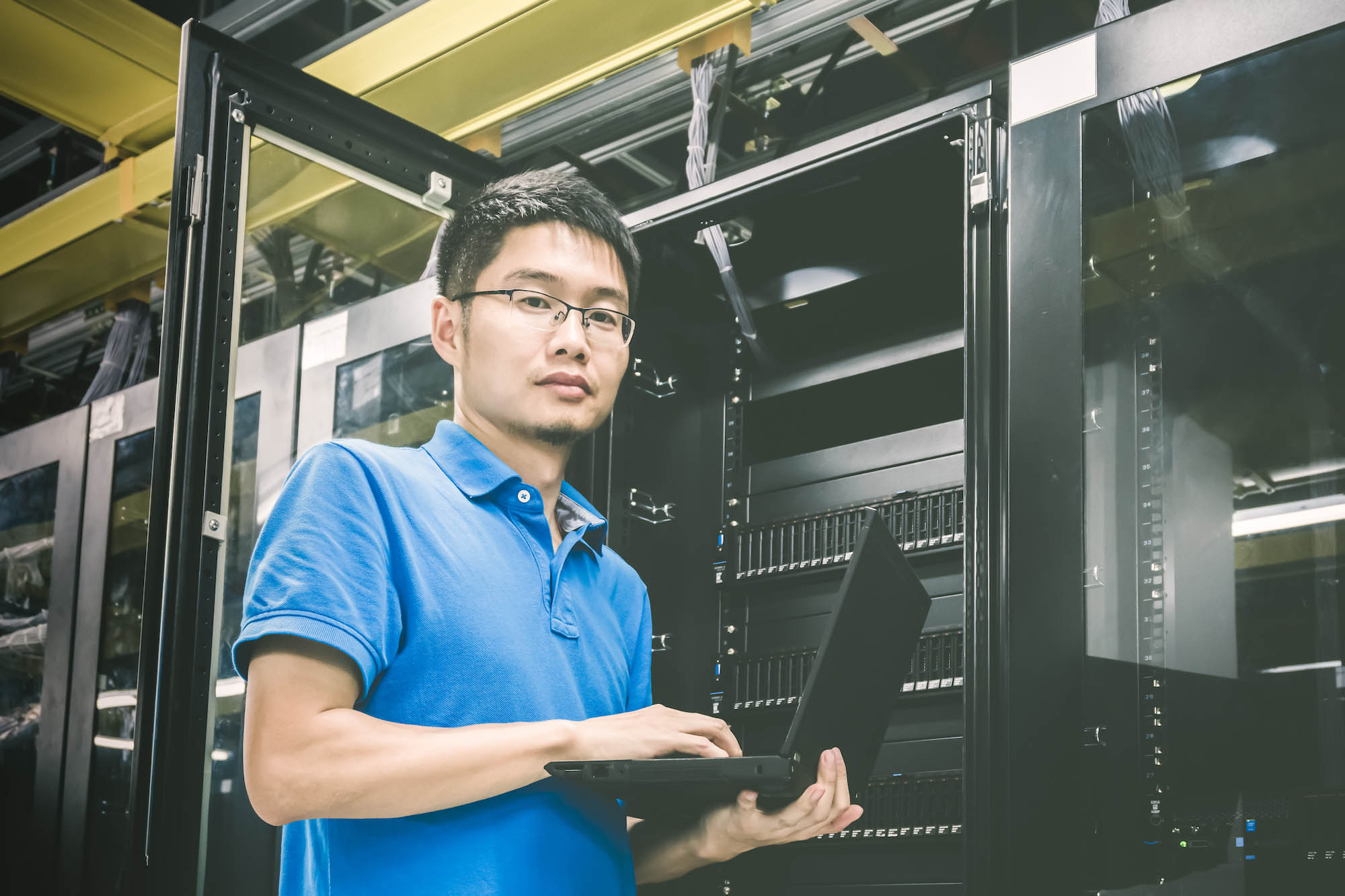 Man in blue shirt using a laptop while standing in front of a server rack.