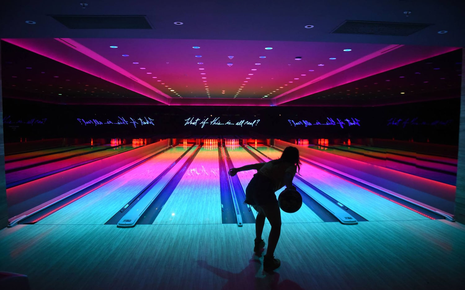 A woman bowling in an alley with vibrant neon lights and glowing lanes, with a text projection asking 