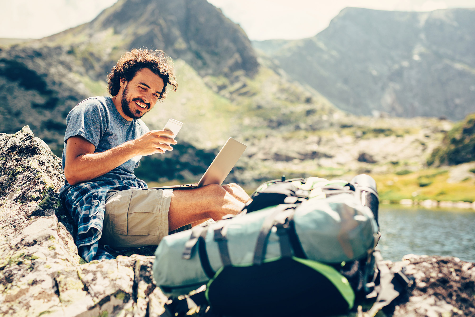 Smiling man with a laptop and smartphone sitting on a rocky mountain.