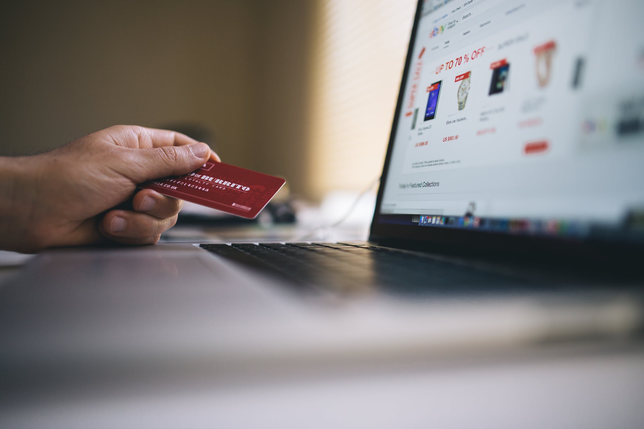 A close-up of a person's hand holding a red credit card next to a laptop with an online shopping site open, indicating an online transaction in progress.