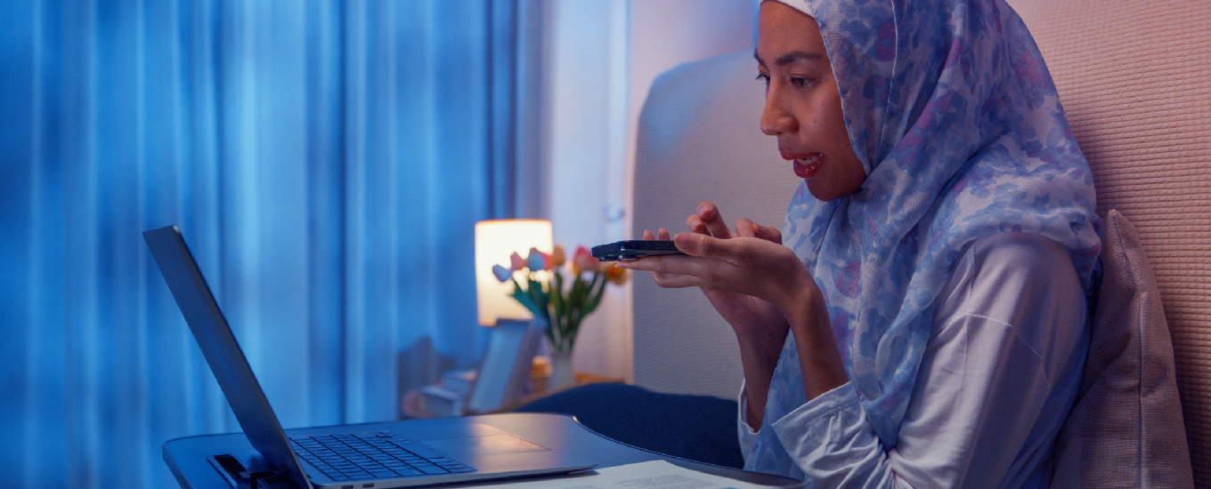 A woman wearing a hijab speaking into a mobile phone while working on a laptop in a home setting.