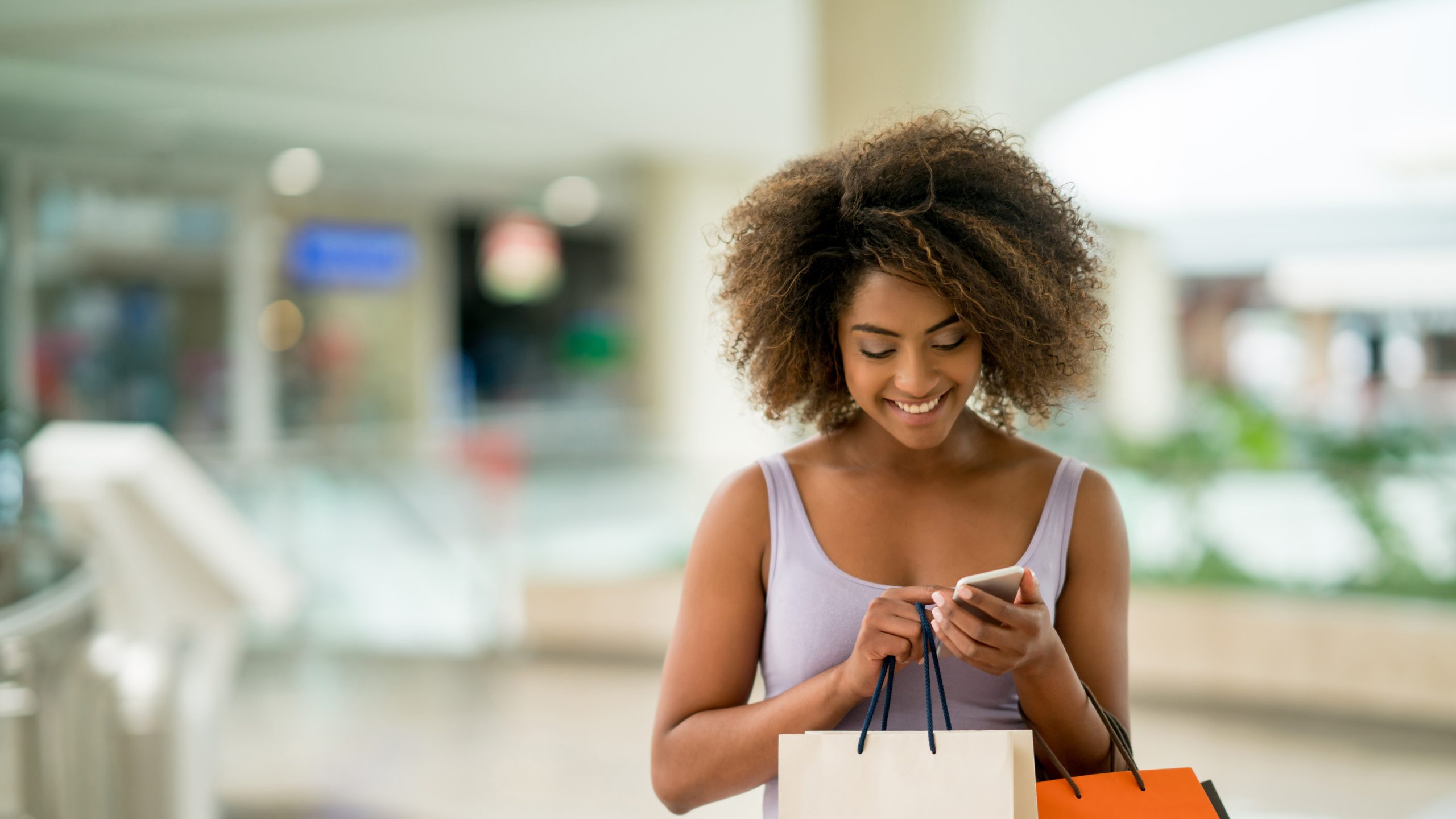 A happy woman with curly hair, holding shopping bags and looking at her phone, standing in a sunlit, modern mall.