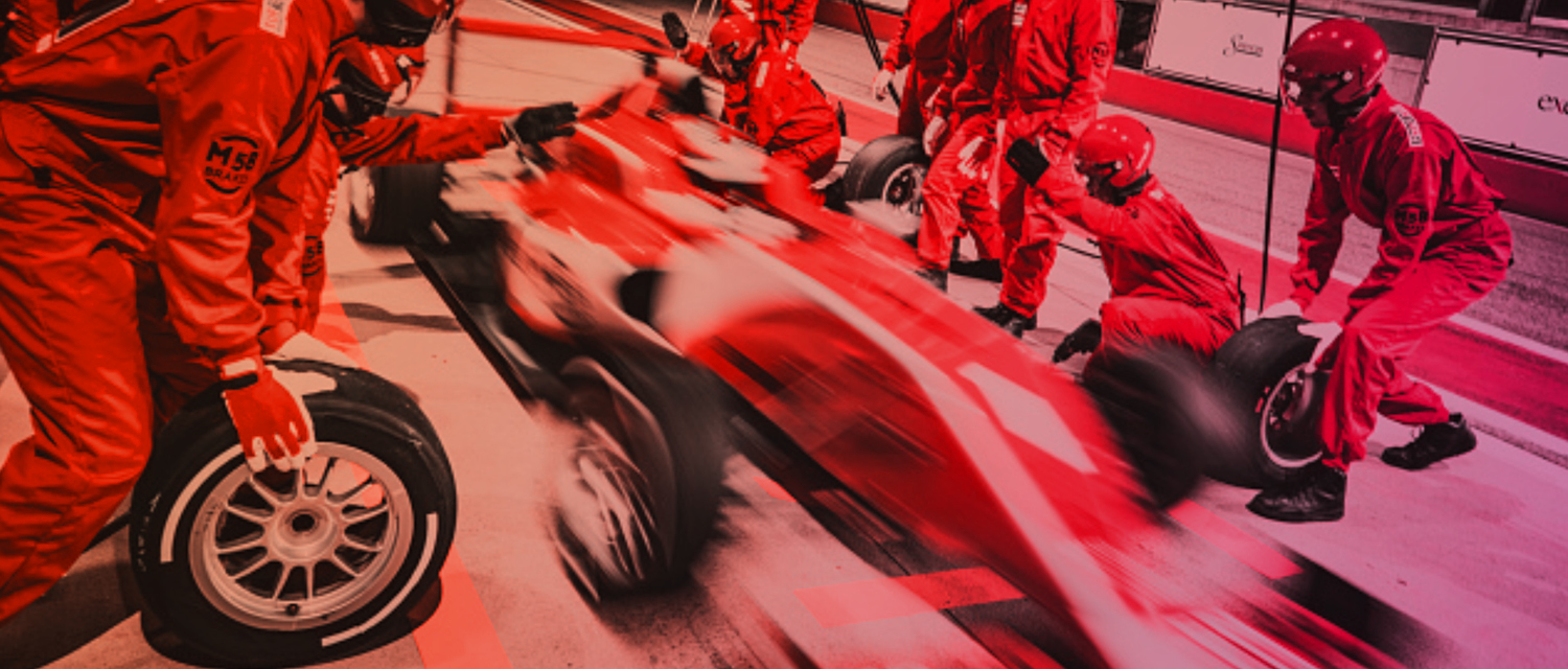 A red-tinted image of a racing pit stop with a team of mechanics in red uniforms changing tires on a blurred race car.