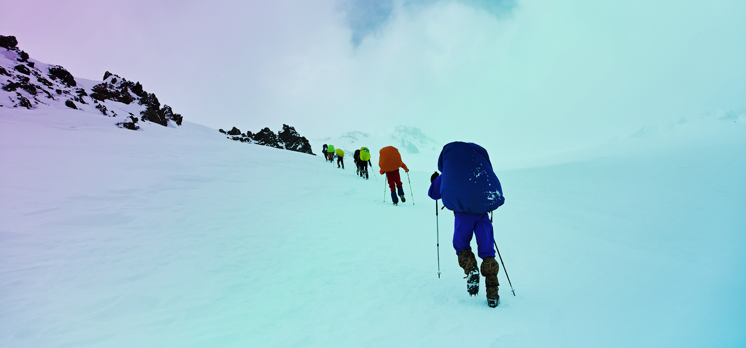 A group of hikers in bright clothing ascending a snow-covered slope in a mountainous environment, under a cloudy sky.