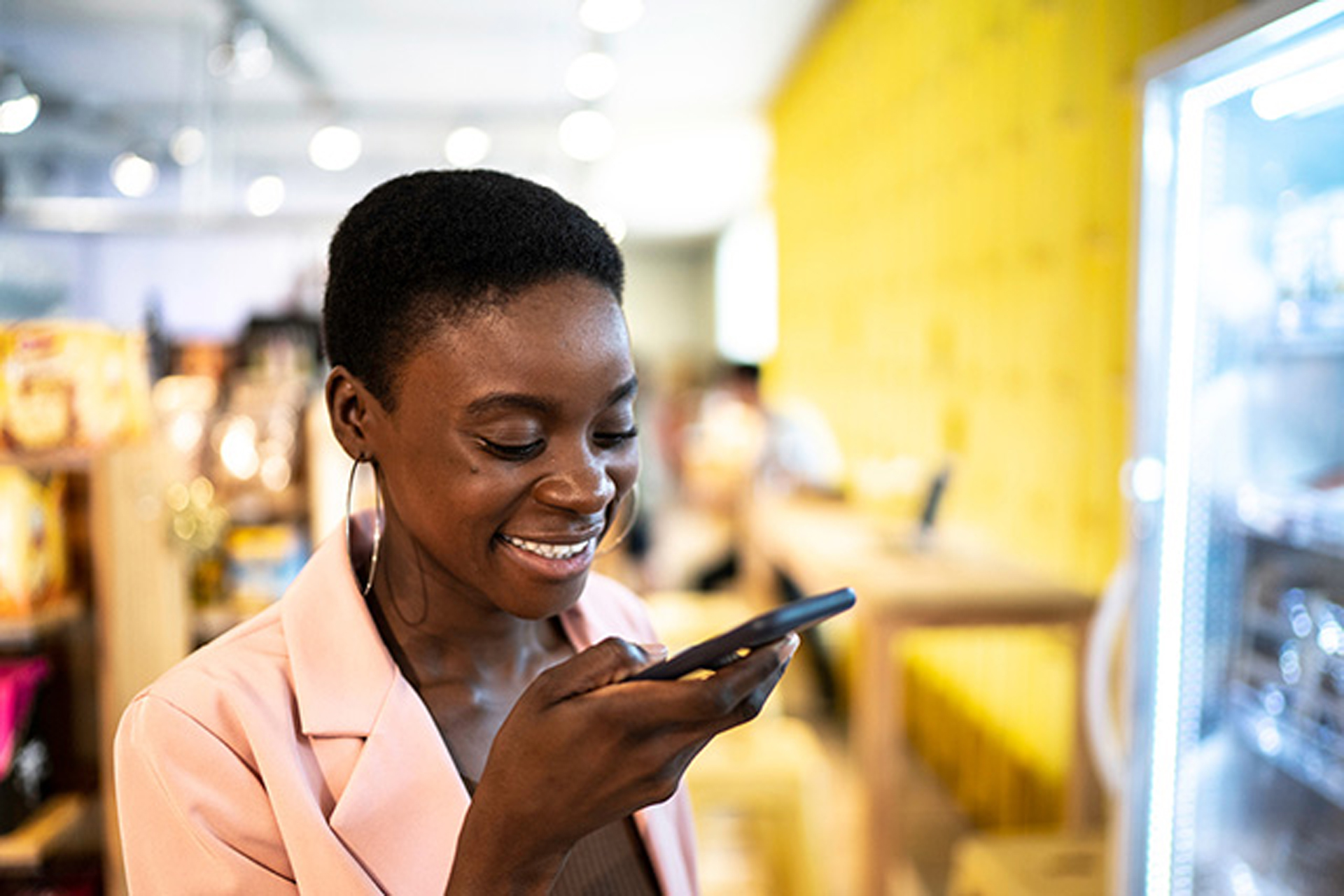 A cheerful woman in a light pink blazer speaking into her phone in a brightly lit store.