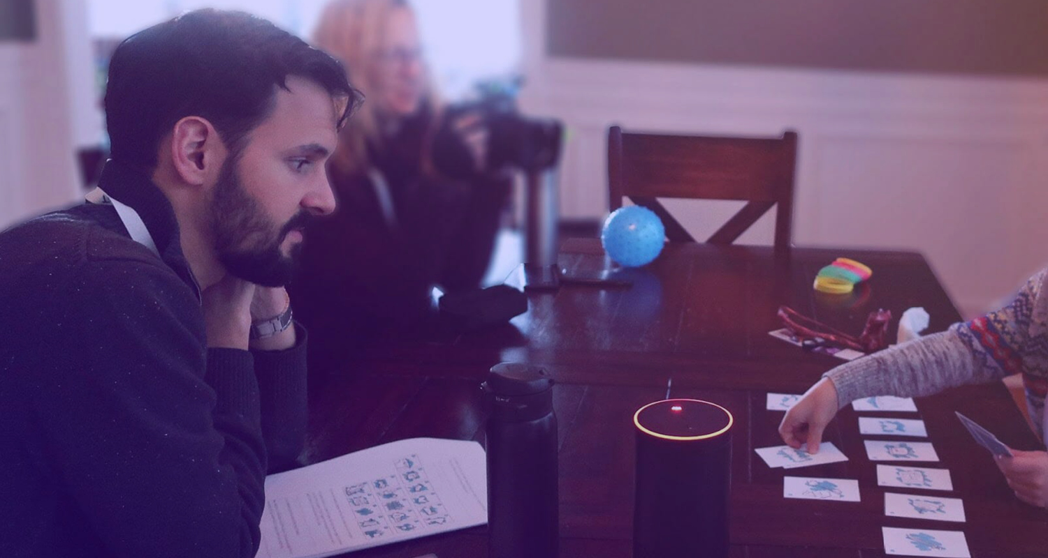 A man seated at a dining table looking thoughtfully at an Alexa voice assistant device while other participants in the background engage with cards and a camera.