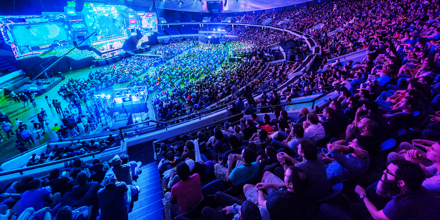A vibrant, high-energy esports arena filled with spectators under blue and purple lights, watching a game on large screens.